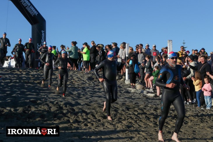 Athletes in wetsuits competing in a triathalon, running on the beach.